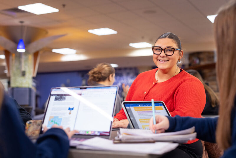 Female student in an orange sweater smiles while studying at a laptop in a campus common area with other students visible in the background