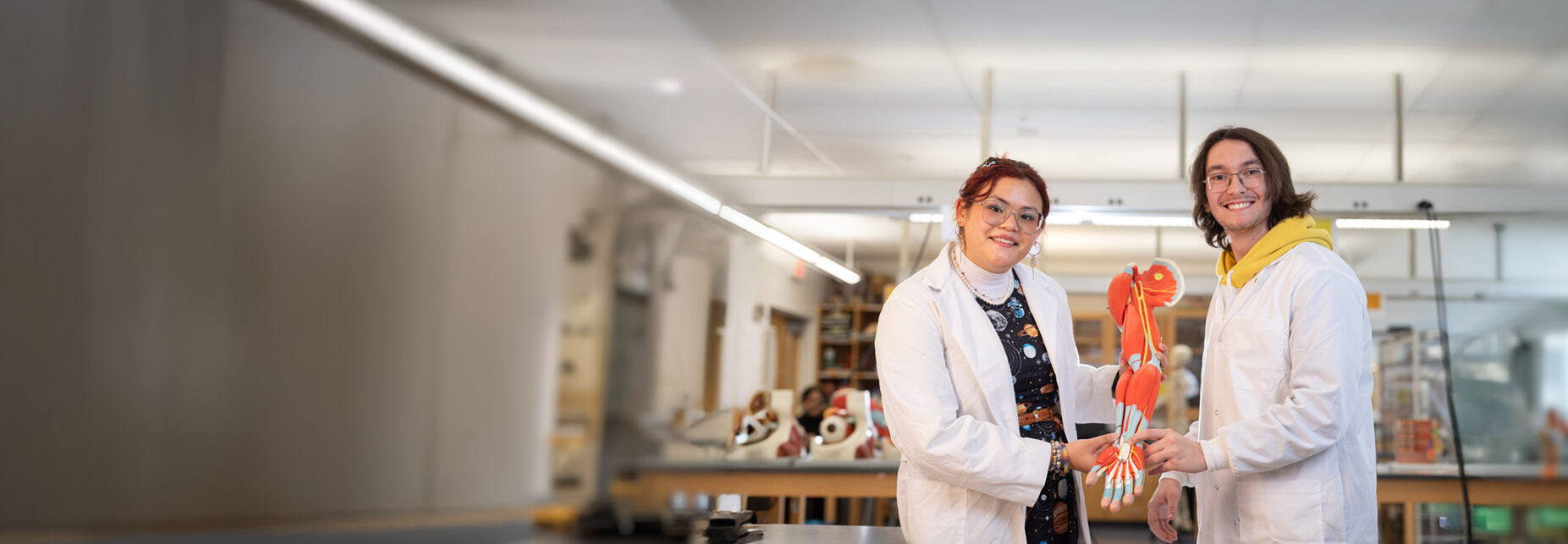 Two students in white lab coats smile while holding an anatomical model of a human arm in a science laboratory